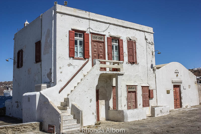 Windmills of Mykonos, Whitewash Buildings: Why We Love this Cruise Port 6 A large white house with red trim on a cobblestone street seen as we walked from the Mykonos cruise port