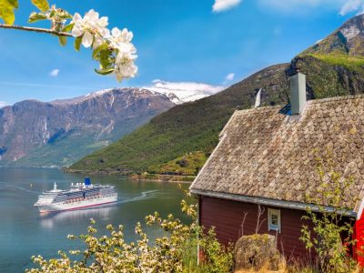Cruise ship sailing into one of the Norway Cruise Ports in a fjord with a red cottage in the foreground