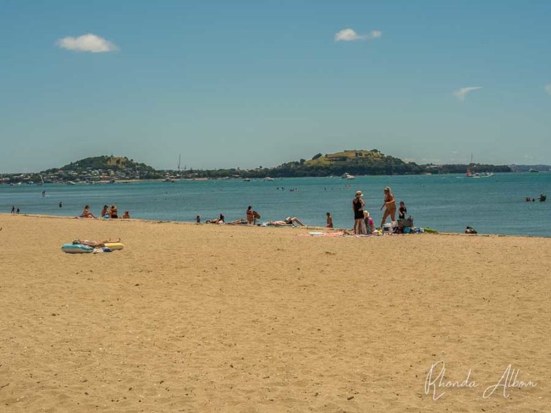 People on Mission Bay beach in Auckland, New Zealand with Devonport seen across the bay